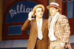Two men on stage dressed as characters from a musical, one in suit and hat, the other in striped jacket and bowler hat,