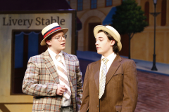Two men on stage in costumes, one wearing a suit and tie, the other in a plaid jacket and hat, engaged in conversation.