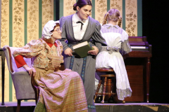 Two women on stage dressed in period costumes, one reading a book to the other who is seated and holding a small child.