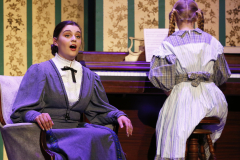 Woman in blue dress sitting at piano, child standing behind her, both looking towards camera.