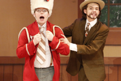 Two people on stage in a musical performance, one dressed as a band member and the other as a conductor, both wearing