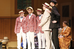 Theatrical performance of a musical production featuring a group of young men in colorful costumes and hats, standing on