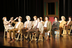 Theatrical stage performance of a musical play featuring actors in period costumes, posing for a group photo.