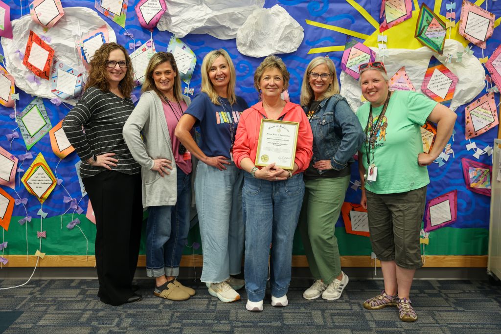 Group of people posing in front of a bulletin board decorated with children's artwork and certificates, smiling and proudly