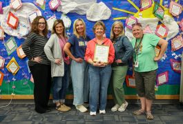Group of people posing in front of a bulletin board decorated with children's artwork and certificates, smiling and proudly