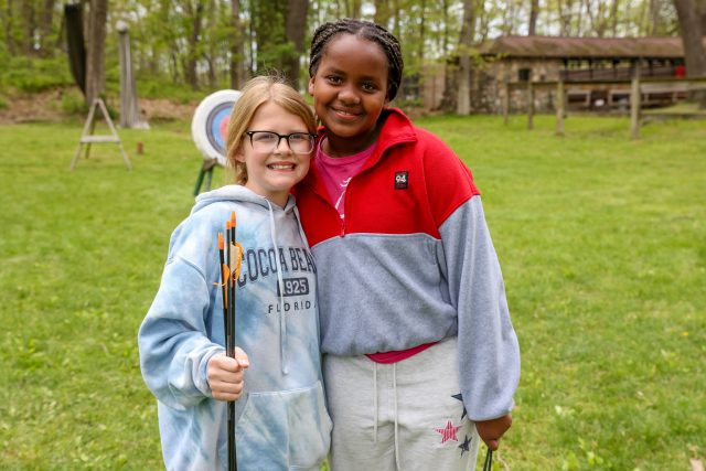 Two people posing for a picture on a grassy field, one holding a bow and arrow.