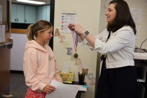 Woman in a white shirt pinning medal on girl's shirt.