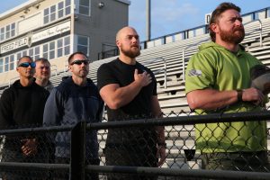 Group of men in stadium stands, some standing, others sitting, all looking towards center where man is speaking into