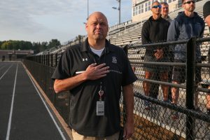 Man standing on a football field, giving the peace sign, wearing a black shirt and khaki pants.