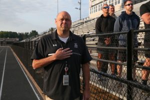 Man standing on bleachers at a sporting event, holding a microphone, speaking to an audience.