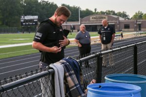 Man in black shirt standing on sideline of football field, talking to man in white shirt.