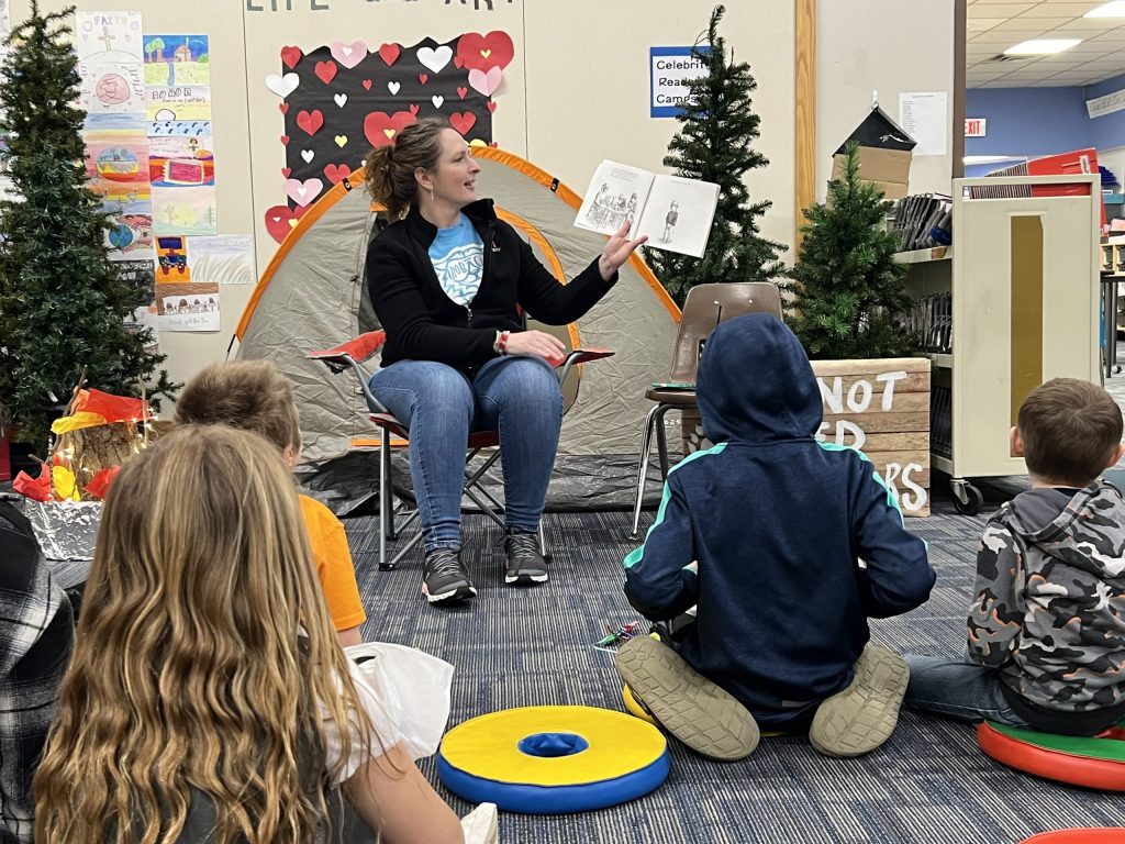 Adult female reading a book to students sitting on the floor in front of her