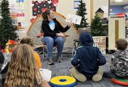 Adult female reading a book to students sitting on the floor in front of her