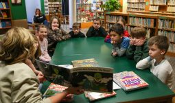 Children gathered around a table in a library setting, listening to an adult reading a book aloud.