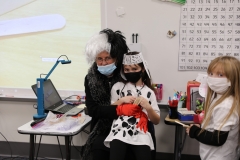 Woman in mask at head of table with children, one holding stuffed dog, all wearing masks and looking at camera, classroom