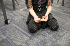 Young girl in a classroom setting, sitting on the floor, holding a small wooden catapult and looking down at it.
