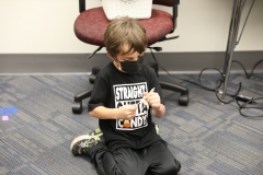 Young boy sitting on floor in front of desk, wearing mask and holding pumpkin catapult.