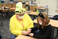 Teacher wearing a face mask and Minnie Mouse ears, sitting at a desk with a child, both engaged in an activity.