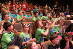 Group of people in a theater, some clapping, others watching, all wearing masks; audience members seated on chairs and