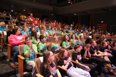 Crowd of people sitting in rows of chairs, clapping their hands in a theater setting.