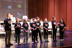 Team of children at a podium, posing for a picture after participating in a spelling bee competition.