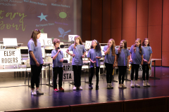 Children on stage in front of microphones, holding signs with names of cities and states, participating in a spelling