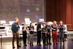 Students on stage at a school event, posing for a picture in front of a sign advertising the 2024 PHM Elementary Spell Bowl