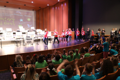 Children on stage performing at a school event, with audience clapping in support.