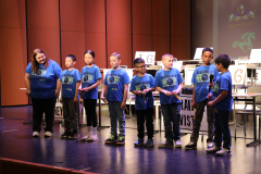 Children in blue shirts standing on stage at a school event, posing for a picture with a sign that reads "2024 PHM