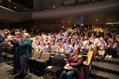 Auditorium filled with people seated in rows of chairs, watching a performance on stage.
