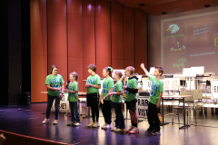 A group of children on stage at a school event, posing in front of a podium and presentation screen.