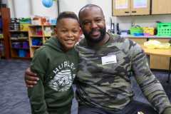 Man wearing camouflage shirt hugging young boy in a classroom setting.