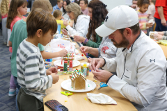 Man in white shirt building gingerbread houses with children at a table.