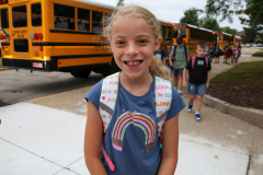 Girl in a blue shirt standing next to a school bus.