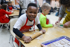 Boy in apron at table making food, smiling, looking at camera.
