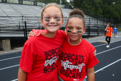 Two young girls posing together in front of a stadium, both wearing red shirts and black shorts, one with glasses and both