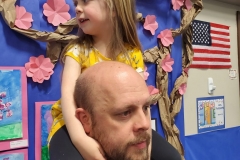 This is a photograph of a man carrying a young girl on his shoulders in front of a backdrop that includes a tree and a sign