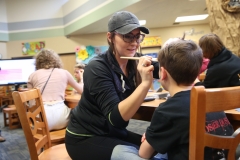 Woman in black shirt applying makeup to boy's face at table in room with chairs and people.