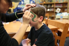 Woman applying green face paint to boy's face in library setting.