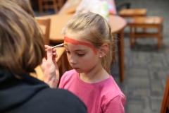 Young girl applying makeup to her face in a room with dining tables and chairs.