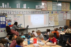 Teacher at front of classroom showing video on screen to students seated at desks.