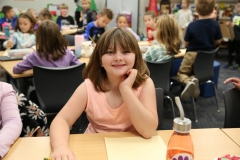 Little girl sitting at a table in a classroom setting, smiling at the camera.