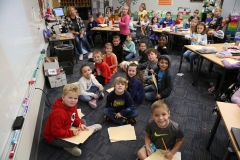 Children sitting at desks in a classroom, some holding pencils, posing for a group picture.