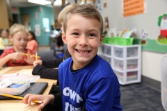 Boy in blue shirt smiling at camera, sitting at desk with pencil, book, and other school supplies behind him.