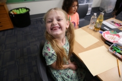 Little girl sitting at a table in a classroom, smiling broadly at the camera.