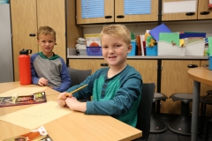 Two boys sitting at a table in a classroom, writing on paper, surrounded by school supplies and chairs.