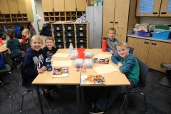 Three children sitting around a table in a classroom, smiling at the camera. The table is cluttered with various items