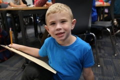 A young boy sitting at a table in a classroom, holding a clipboard and looking directly at the camera.