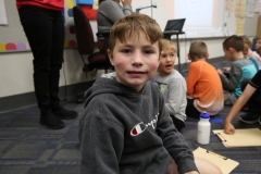 Boy in grey hoodie sitting on floor next to table with paperwork, smiling at camera.