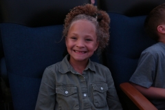 Little girl in a theater seat, smiling at camera.